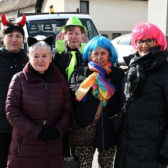 Fünf Frauen stehen draußen, alle in farbenfrohen, skurrilen Kostümen gekleidet. Sie tragen verschiedene Accessoires wie Hüte, Perücken und Schals. Hinter ihnen ist ein weißer Van und ein Gebäude zu sehen.