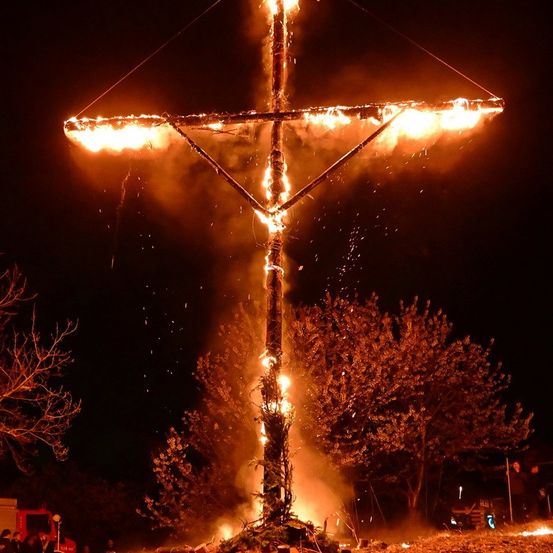 Ein großes Kreuz brennt bei Nacht. Das Feuer ist so intensiv, dass es die gesamte Umgebung beleuchtet. Bäume umgeben das Kreuz, und es stehen Leute in der Nähe.