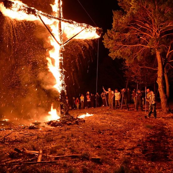 Ein großes Lagerfeuer mit einem brennenden Kreuz, umgeben von Menschen mit Stöcken, bei Nacht.