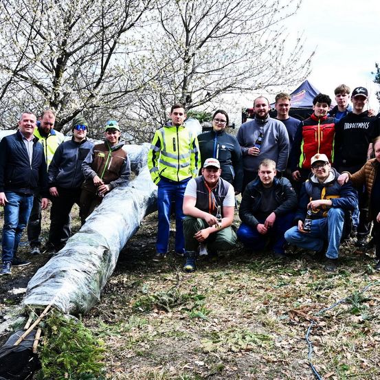 Eine Gruppe von Personen posiert im Freien für ein Foto, mit einem großen Baumstamm in der Mitte. Einige stehen, während andere sitzen. Bäume und ein Zelt sind im Hintergrund zu sehen.