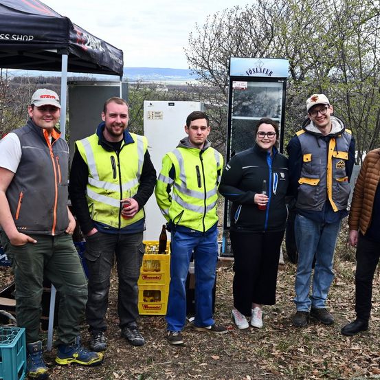 Eine Gruppe von Personen in Warnwesten steht auf einem Feld. Einige halten Getränke. Ein Zelt und Kisten befinden sich im Hintergrund. Bäume und ein Schild sind in der Ferne.