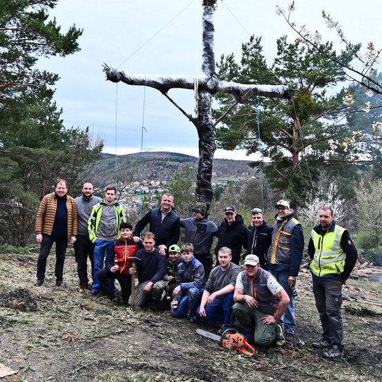 Eine Gruppe von Männern posiert mit einem hölzernen Kreuz auf einem Hügel. Einige halten Werkzeuge, und einer kniet mit einer Kettensäge. Bäume und Berge sind im Hintergrund.