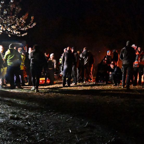 Eine Gruppe von Menschen ist im Dunkeln um ein Feuer versammelt, einige sitzen, andere stehen. Eine Frau hält ein Baby und ein Mann kniet. Es gibt einen Baum mit Blumen und ein Fahrzeug in der Nähe.