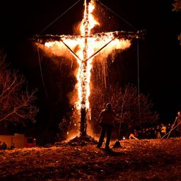 Ein loderndes Lagerfeuer mit einem Kreuz auf der Spitze brennt in der Nacht, mit Menschen um es herum. Eine Person hält eine Besen. Bäume und ein rotes Gebäude sind im Hintergrund.