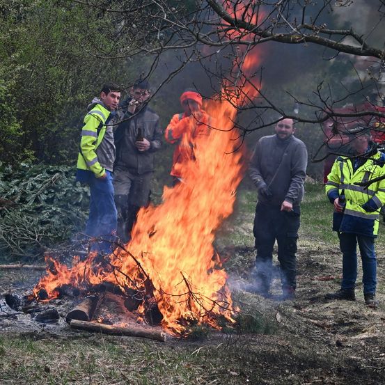 Mehrere Männer beobachten ein brennendes Feuer auf einem Feld. Ein Mann hält ein Bier.
