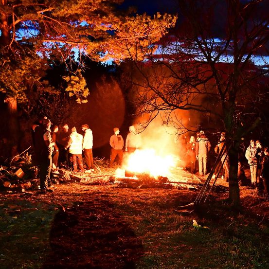 Eine Gruppe von Menschen ist um ein Lagerfeuer in einem Wald bei Nacht versammelt. Sie tragen Winterkleidung, einige stehen und andere sitzen. Bäume umgeben den Bereich, und das Feuer brennt hell.