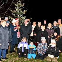 Eine Gruppe von Menschen steht bei Nacht um einen Weihnachtsbaum, einige tragen Winterkleidung und Mützen. Einige knien auf dem Gras.