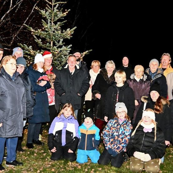 Eine Gruppe von Menschen steht bei Nacht um einen Weihnachtsbaum, einige tragen Winterkleidung und Mützen. Einige knien auf dem Gras.