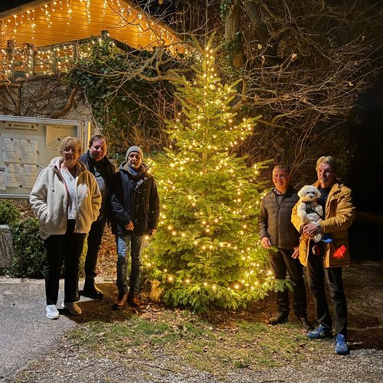 Fünf Personen posieren für ein Foto neben einem beleuchteten Weihnachtsbaum, in Winterkleidung, vor einem beleuchteten Haus.
