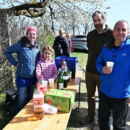 Vier Menschen, zwei Männer und zwei Frauen, stehen vor einem Tisch mit Getränken. Ein junges Mädchen steht zwischen den Frauen. Auf dem Tisch stehen Weinflaschen und eine Kiste mit Pfirsichen. Ein Auto und ein Baum sind im Hintergrund zu sehen.