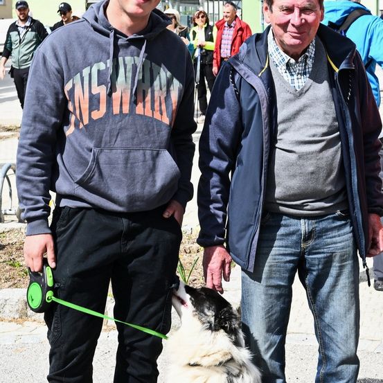 Ein Mann und ein älterer Mann stehen mit einem Hund an der Leine und lächeln in einer Außenumgebung, mit Menschen im Hintergrund, die vorbeigehen.