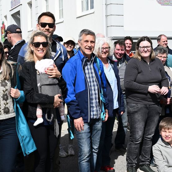 Eine Gruppe von Menschen steht draußen vor einem Gebäude. Eine Frau hält ein Baby in den Armen. Der Mann in der Mitte lächelt, während andere hinter ihm stehen.