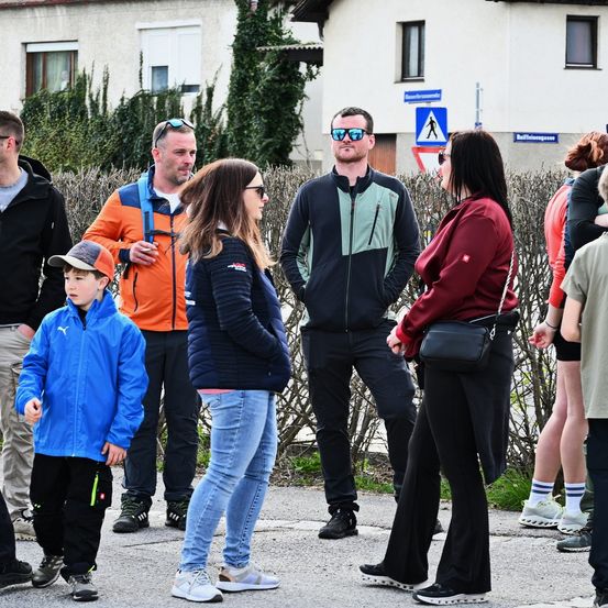 Eine Gruppe von Menschen steht draußen. Einige tragen Brillen, Mützen und Turnschuhe. Der Junge trägt eine blaue Jacke. Der Mann mit Sonnenbrille steht hinter der Frau, die eine rote Jacke trägt. Sie stehen vor einem Gebäude.