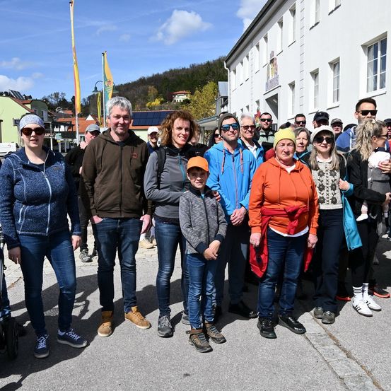 Eine Gruppe von Menschen steht vor einem weißen Gebäude mit vielen Fenstern. Einige tragen Hüte und Brillen. Der Junge in der Mitte trägt einen orangefarbenen Hut. Bäume und Häuser befinden sich hinter ihnen.