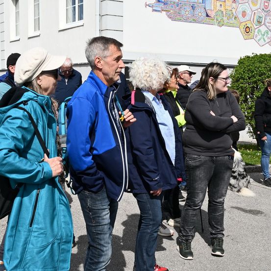 Mehrere Erwachsene, einige mit Mützen und Brillen, stehen vor einem Gebäude mit einem Wandbild. Sie sind in verschiedenen Farben gekleidet und einige tragen Rucksäcke.