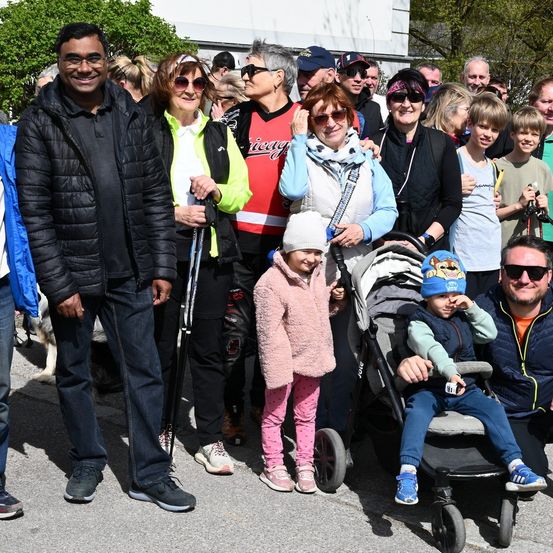 Eine vielfältige Gruppe von Menschen, einschließlich Erwachsener und Kindern, posiert für ein Foto im Freien. Einige halten Skistöcke, während ein Kind in einem Kinderwagen sitzt. Bäume und ein Gebäude sind im Hintergrund.