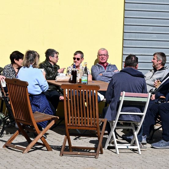 Eine Gruppe von Menschen sitzt draußen an einem Tisch. Auf dem Tisch befinden sich Flaschen und andere Gegenstände. Sie schauen alle in verschiedene Richtungen.