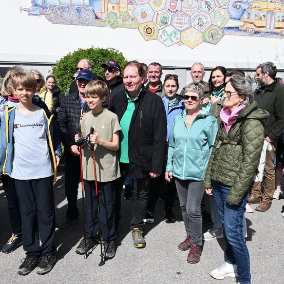 Eine Gruppe von Menschen, darunter Kinder, steht vor einer Wand mit einem Mosaik-Design. Einige tragen Brillen und andere halten Trekkingstöcke. Sie scheinen für ein Foto zu posieren.