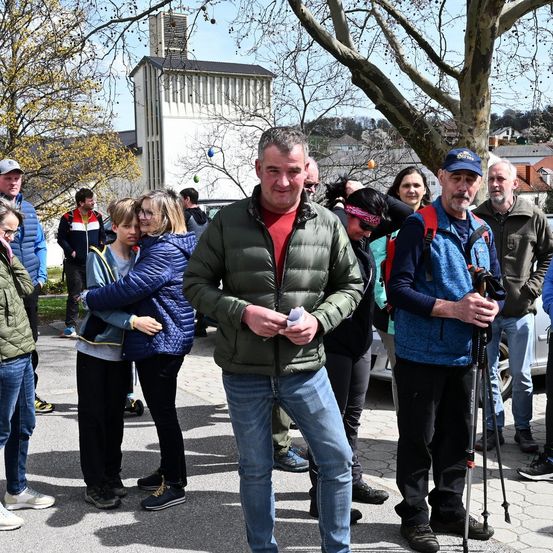 Eine Gruppe von Menschen, einige in Jacken und andere in Mützen, versammelt sich um einen Mann, der ein Stück Papier in der Hand hält, mit einem Gebäude und Bäumen im Hintergrund.
