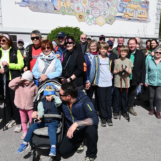 Eine Gruppe von Menschen, einschließlich Kindern und Erwachsenen, versammelt sich vor einer Wand mit einem Zug- und Busbild. Einige halten Stöcke.