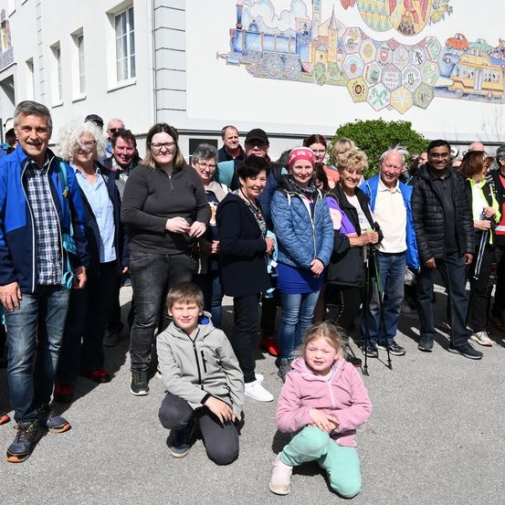 Eine Gruppe von Menschen posiert für ein Foto vor einem Gebäude mit einem Wandbild. Ein Junge und ein Mädchen knien davor.