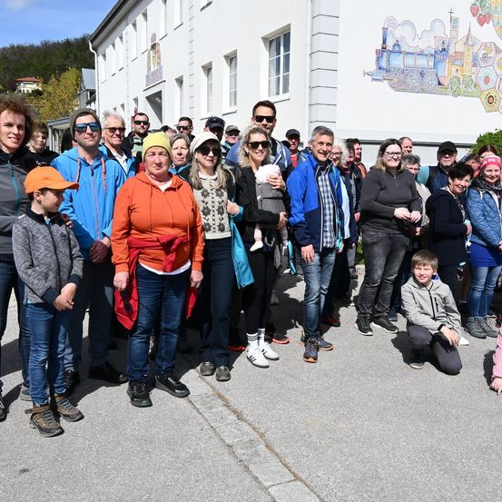 Eine Gruppe von Menschen posiert für ein Foto vor einem Gebäude. Einige tragen Sonnenbrillen und Mützen. Eine Frau hält ein Baby.
