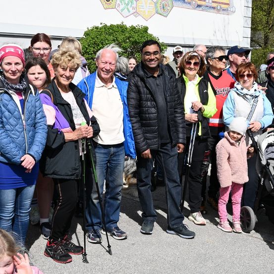 Eine Gruppe von Menschen in Winterkleidung steht zusammen und lächelt für ein Foto. Sie halten Trekkingstöcke und einige tragen Brillen und Mützen. Ein Baby im Kinderwagen ist in der Nähe und ein Mann trägt einen Rucksack.
