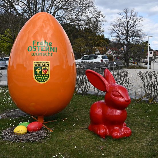 Ein riesiges orangefarbenes Osterei steht neben einer roten Hasenstatue auf einem Rasen. Das Ei hat einen Text, der 'Frohe Ostern! Wunscht' sagt. In der Nähe sind einige Eier in einem Nest.