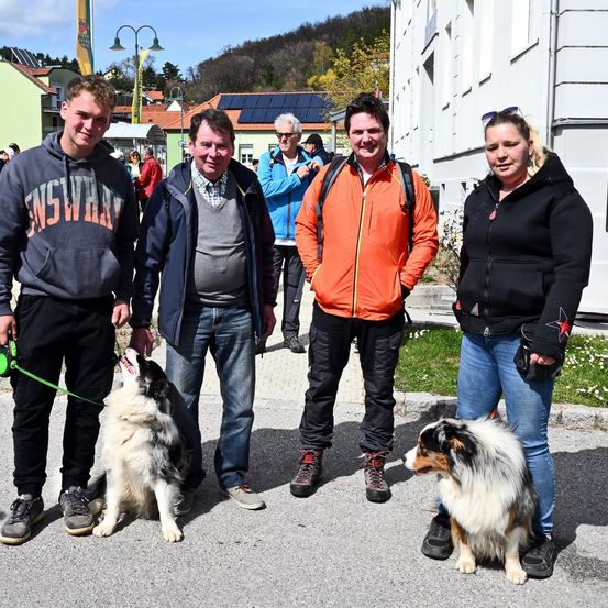 Eine Gruppe von Menschen mit zwei Hunden steht vor einem Gebäude mit Solarpanelen, ein Mann trägt einen Rucksack, ein anderer eine orange Jacke und zwei Frauen schwarze Jacken.