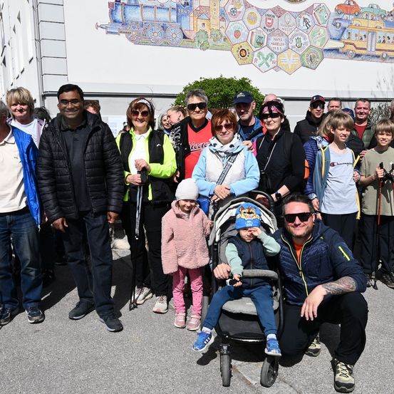 Eine Gruppe von Menschen in Winterkleidung, einige mit Wanderstöcken, posiert vor einer Wand mit einem Wandbild eines Zuges und anderen Bildern.