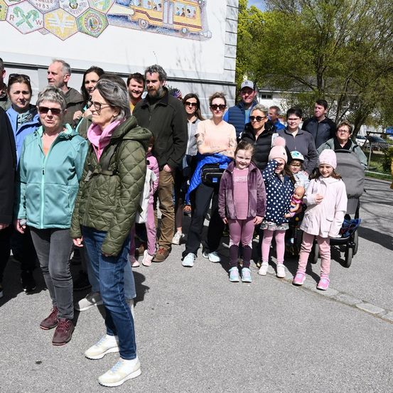 Eine Gruppe von Menschen, darunter Erwachsene und Kinder, steht vor einer Wandmalerei. Einige tragen Brillen und Mützen. Kinder tragen rosa Hosen und halten ein Baby. Ein Kinderwagen steht in der Nähe.