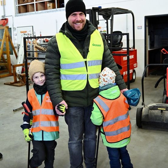 Ein Mann und zwei Kinder in Warnwesten stehen in einem Lager mit Maschinen im Hintergrund.