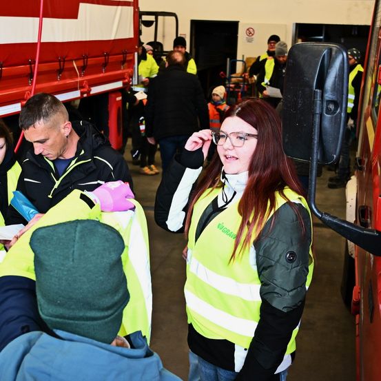 Eine Frau mit roten Haaren in einer Warnweste spricht mit einem Mann mit einer grünen Mütze. Dahinter sind Leute in Warnwesten beschäftigt. Im Hintergrund steht ein Feuerwehrwagen.