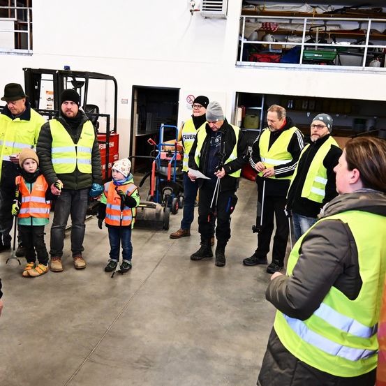Eine Gruppe von Erwachsenen und Kindern in Warnwesten versammelt sich in einem Lagerhaus. Ein Mann hält ein Papier, und ein Kind hält einen Stock.