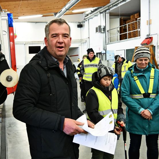 Ein Mann hält ein Papier in der Hand und spricht zu einer Gruppe von Menschen, darunter Kinder, in einem Lager mit Sicherheitswesten.