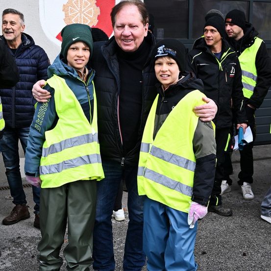 Eine Gruppe von Menschen steht vor einer Wand mit einem Logo. Zwei Jungen in reflektierenden Westen und Mützen stehen neben einem Mann in einer schwarzen Jacke. Dahinter stehen ein Mann mit einer Mütze und eine Person in einem weißen Hemd.