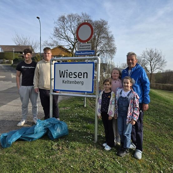 Eine Familie steht neben einem Wiesen Keltenberg-Schild in einem Grasbereich. Sie alle lächeln und posieren für ein Foto.