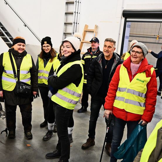 Eine Gruppe von Menschen in Warnwesten steht in einem Gebäude, einer hält einen Stock. Dahinter steht eine Person in der Nähe einer Tür.