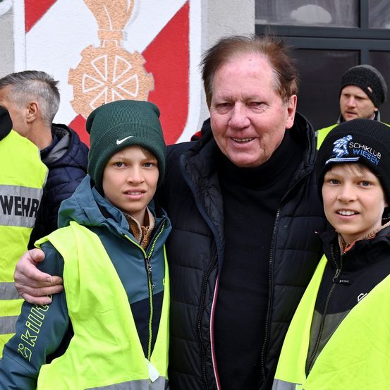 Ein Mann in einem schwarzen Jackett lächelt für ein Foto mit zwei Kindern in neonfarbenen Westen. Sie stehen vor einer Wand mit einem roten und weißen Design.