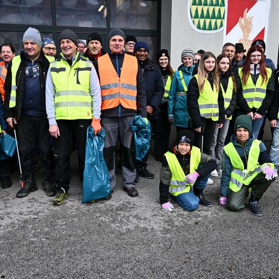 Eine Gruppe von Menschen in Warnwesten und Mützen steht vor einem Gebäude, einige halten Plastiktüten. Sie scheinen für ein Foto zu posieren.