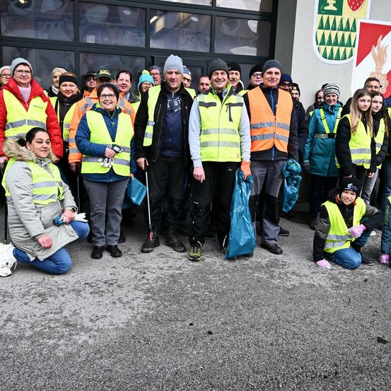 Eine Gruppe von Menschen in Reflektorwesten, einschließlich Männer und Frauen, posiert für ein Foto vor einem Gebäude mit einem Wappen an der Wand. Einige knien und einige stehen. Sie halten blaue Taschen.