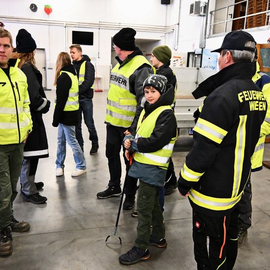 Eine Gruppe von Menschen in gelben Warnwesten steht in einem Raum mit einem Kind, während ein Feuerwehrmann mit ihnen spricht.