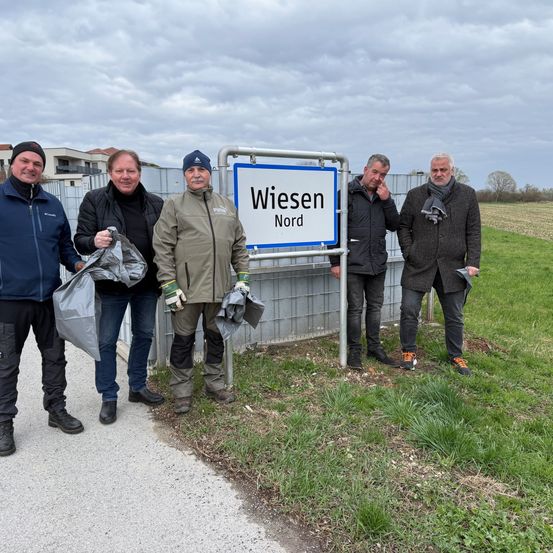 Fünf Männer stehen neben einem Straßenschild für Wiesen Nord und posieren für ein Foto. Sie halten Plastiktüten in der Hand und scheinen an einer Gemeinschaftsaktivität beteiligt zu sein.