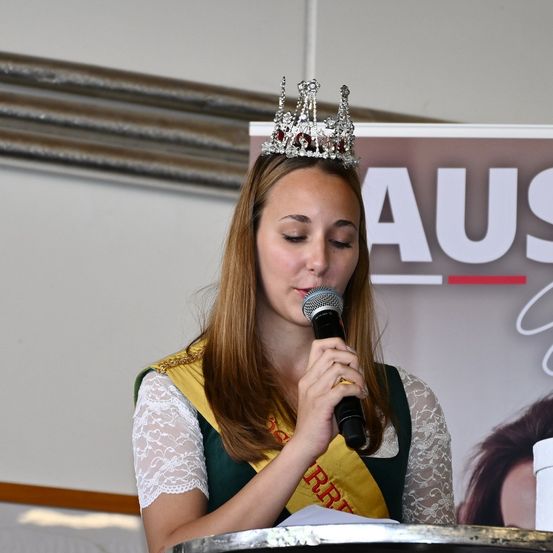 Eine Frau mit Krone hält ein Mikrofon und spricht. Sie trägt einen grün-gelben Schal und ein weißes Spitzenkleid. Hinter ihr befindet sich ein Schild mit dem Wort 'AUS'.