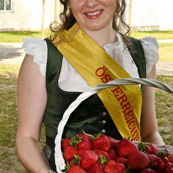 Eine Frau lächelt und hält einen Korb voller Erdbeeren. Sie trägt eine gelbe Schärpe und ein grünes Kleid mit weißen Abschlüssen. Der Korb ist voll mit frischen roten Erdbeeren.
