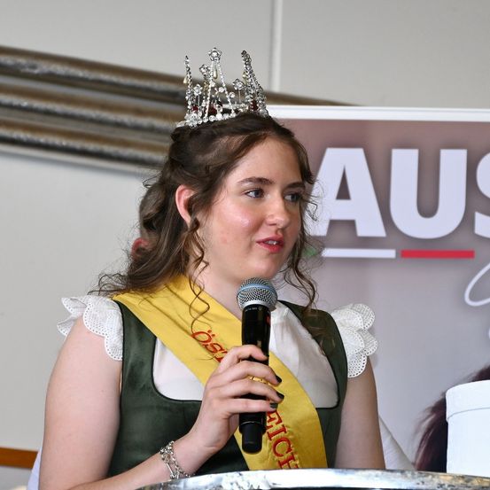 Eine Frau mit Krone und Mikrofon spricht bei einer Veranstaltung. Sie trägt ein grünes und gelbes Band. Hinter ihr ist ein Banner mit der Aufschrift 'AUS' zu sehen.