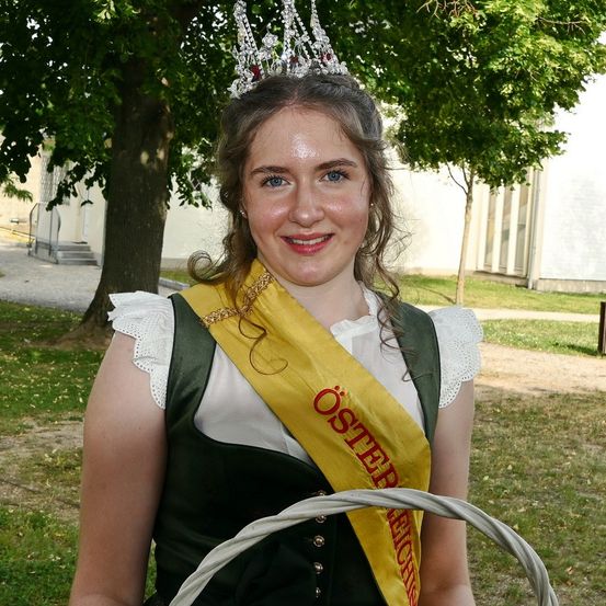 Eine Frau in traditioneller Kleidung hält einen Korb und trägt eine Krone. Sie steht in einem Garten mit Bäumen und einem Gebäude dahinter.