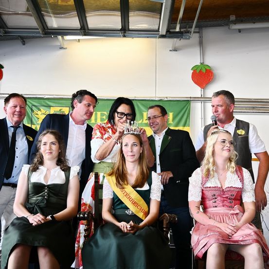 Eine Gruppe von Menschen in traditioneller deutscher Kleidung versammelt sich für ein Foto. Eine Frau wird mit einer Tiara gekrönt. Hinter ihnen sind Banner und eine Erdbeere zu sehen. Ein Mann mit Mikrofon steht in der Nähe.