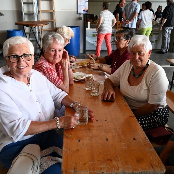 Eine Gruppe von Frauen sitzt gemeinsam an einem Holztisch. Gläser und Teller mit Essen stehen vor ihnen. Andere Personen stehen im Hintergrund.