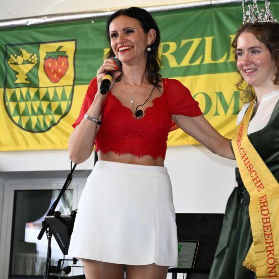 Eine Frau mit Mikrofon steht neben einer Frau mit Krone. Sie lächeln beide und hinter ihnen ist ein Banner mit einem Logo und Text.